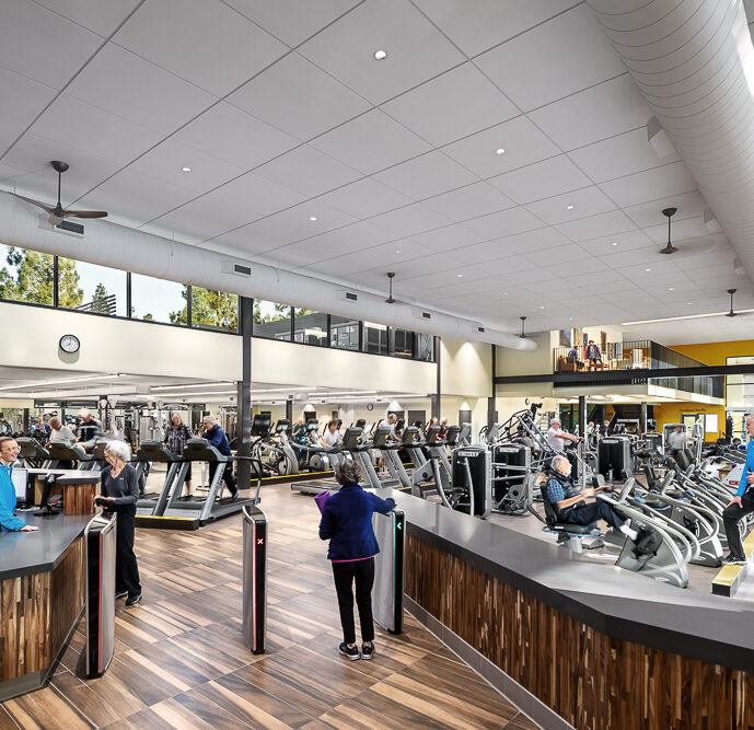 the interior of a modern fitness center with people exercising on treadmills and other cardio equipment. The reception area features a wooden desk where a staff member is assisting a gym-goer, while another person in blue enters the space. Natural light streams in through the large windows, and an overhead skylight complements the spacious and airy atmosphere of the gym.