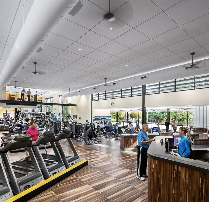 The interior of a modern fitness center with people exercising on treadmills and other cardio equipment. The reception area features a wooden desk where a staff member is assisting a gym-goer, while another person in blue enters the space. Natural light streams in through the large windows, and an overhead skylight complements the spacious and airy atmosphere of the gym.