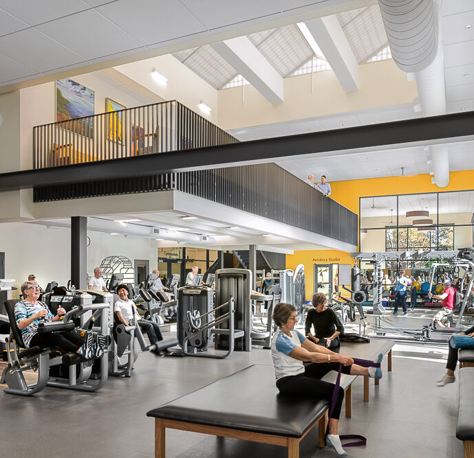 A lively scene inside a fitness center where several people are engaged in various exercises using gym equipment like stationary bikes and weight machines. In the foreground, individuals are seated on benches, possibly in preparation for a workout or cooling down afterwards. The facility has a two-story design with an upper walkway, and its bright interior is enhanced by abundant natural light coming in through the large windows.