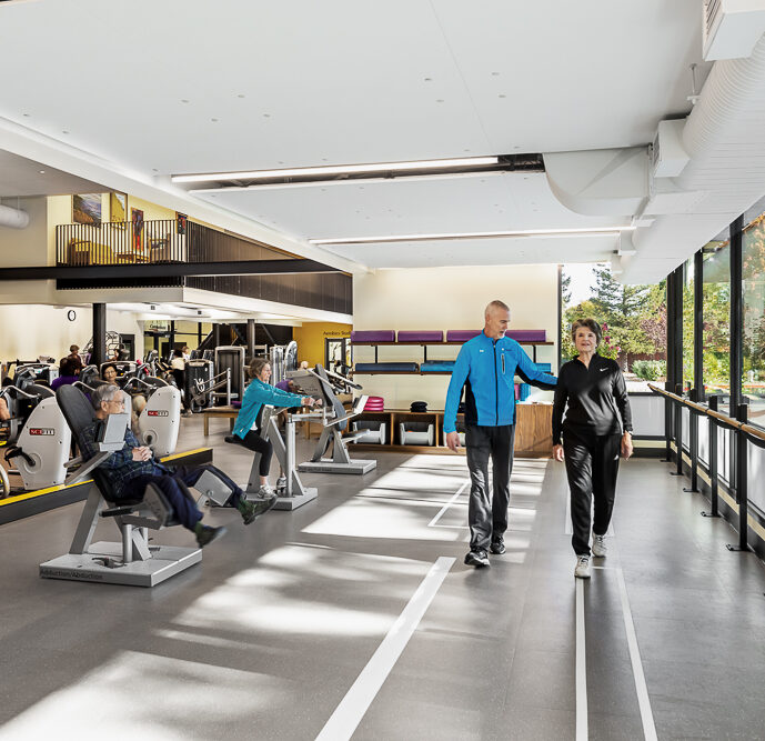 A personal trainer and resident walking through a bright and spacious gym with large windows, while others exercise on stationary bikes and rowing machines. The gym features modern equipment and is designed with a clear walkway marked by white lines on the floor. An upper level with a balcony overlooks the workout area, suggesting a multi-level fitness facility.