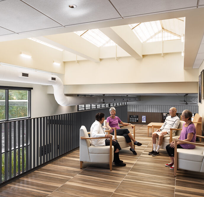 A group of older adults seated and conversing in a comfortable lounge area with modern furnishings and large windows that offer a view of the outside greenery. The interior is well-lit with natural light supplemented by ceiling fixtures, and the walls are adorned with colorful abstract paintings. The space, featuring a high vaulted ceiling and a mix of wood and neutral tones, creates a welcoming and relaxed atmosphere.