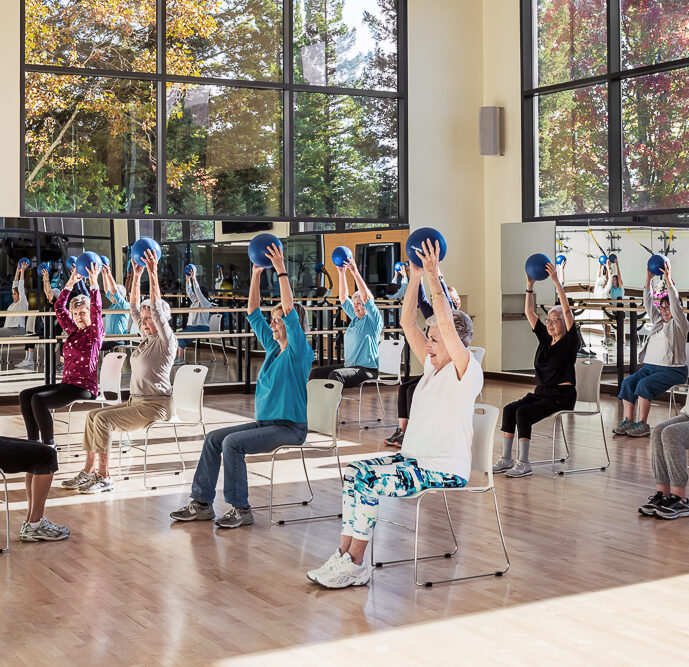 A group of adults participating in a fitness class. They are inside a spacious room with large windows that allow plenty of natural light and offer a view of trees with autumn leaves. The participants are seated on chairs, arranged in rows, and each person is holding a blue exercise ball above their head with both hands. They are wearing casual, comfortable clothing suitable for physical activity. The atmosphere seems to be one of active engagement and communal exercise. The room is equipped with mirrors along the wall, reflecting the participants, which is common in fitness studios to help with form and posture during exercises.