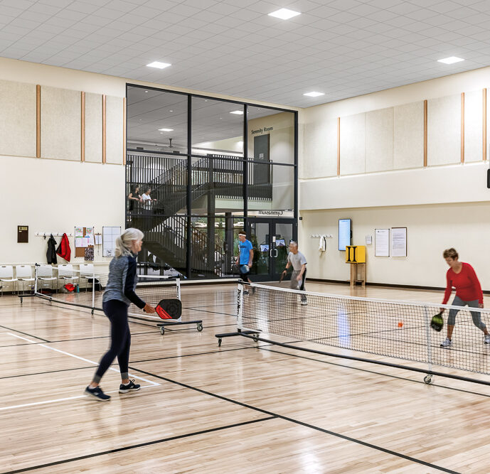 An indoor pickleball match with four participants, two on each side of the net, in a gym with wooden flooring. Spectators can be seen on an overlooking balcony. The players, dressed in athletic wear, are actively engaged in the game.